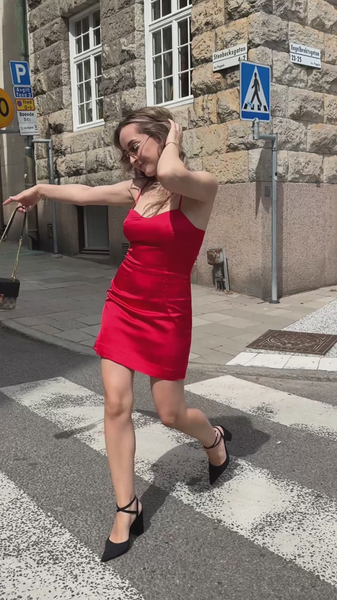 Woman wearing The Gohar red satin mini dress by Leochèle walking across a city crosswalk, smiling and turning toward the camera